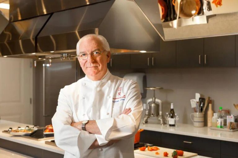 An older male chef with glasses and short white hair stands confidently in a modern Washington kitchen. He wears a white chef’s coat, hinting at his gastronomic expertise, and smiles slightly amid sleek cabinets and stainless steel appliances.