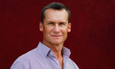 A middle-aged man with short brown hair and blue eyes, wearing a light purple checkered button-up shirt, stands in front of a dark red background, looking confidently at the camera with a slight smile—like an author awaiting the prix Goncourt in littérature française.