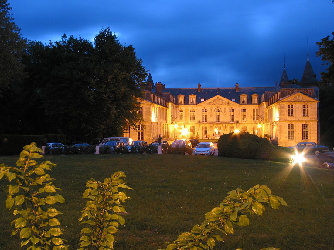 Un grand château bien éclairé évoque le charme des Hôtels Particuliers, avec ses grandes fenêtres et son architecture élégante. Les voitures sont garées devant, tandis que les logements de luxe sont encadrés par des buissons et des arbres jaune-vert sous le ciel crépusculaire. 