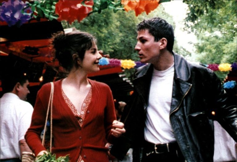 A woman in a red cardigan and a man in a black leather jacket walk hand in hand at an outdoor Paris market decorated with colorful paper flowers, smiling at each other amidst the greenery and lively crowd, enjoying their rendez-vous.