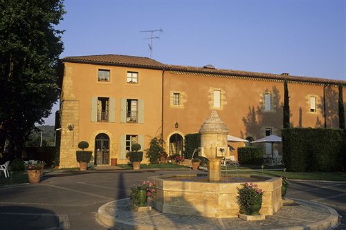 A rustic Perignane bastide with ochre walls, blue shutters, and arched doorways stands behind a round stone fountain. Potted plants, topiary trees, white patio umbrellas, and chairs enhance the courtyard under a clear sky.