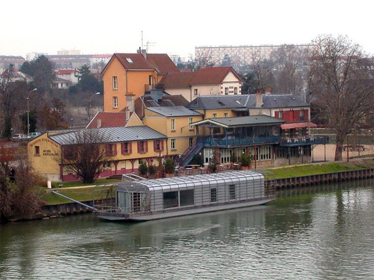 A metal-roofed houseboat is moored on a calm river, reminiscent of Renoir’s Seine scenes. Colorful buildings, including a yellow and red restaurant with outdoor seating, line the shore beneath a cloudy late autumn sky. Apartment blocks rise in the distance.