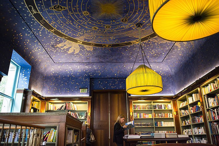 A woman browses books in a cozy library with wooden shelves and tables. The ceiling, painted dark blue with gold stars, evokes littérature Française, while two large yellow lamps create a warm, inviting atmosphere reminiscent of a marché Américain.
