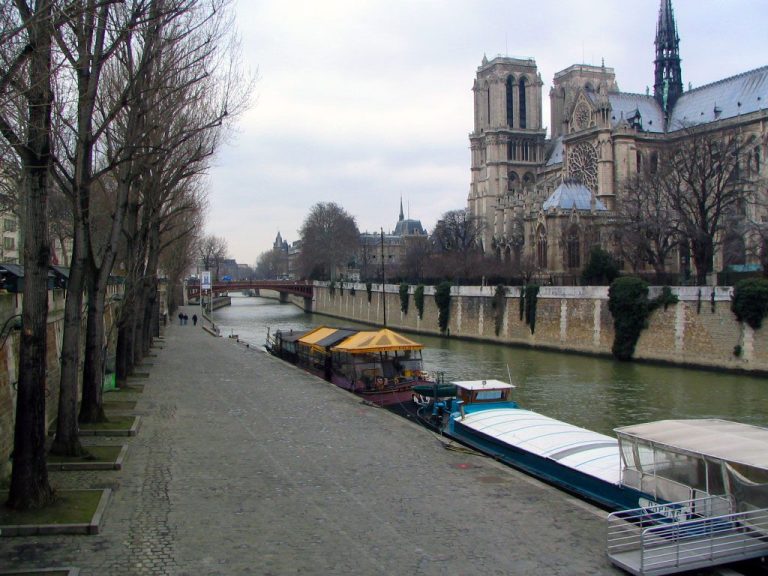 Vue de la Seine à Paris avec des péniches amarrées le long des berges, des arbres dénudés bordant une allée pavée et l'architecture gothique de la cathédrale Notre-Dame sur la droite, le tout au milieu des sons charmants de Paris sous un ciel nuageux.