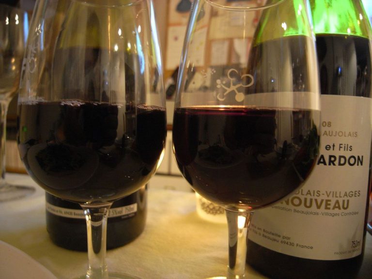 Two glasses of red wine on a table, positioned in front of two bottles—one showcasing the arrival of Beaujolais Nouveau. The softly blurred background with pinned menus creates a cozy ambiance, while light dances off the vin français-filled glasses.