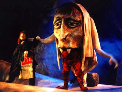 A person in a cloak stands beside a performer wearing a large, exaggerated troll marionnette with fangs and a big nose. The stage, set with rocky props and blue lighting, evokes the mystical world of La Couronne du Destin and its legendary géants.