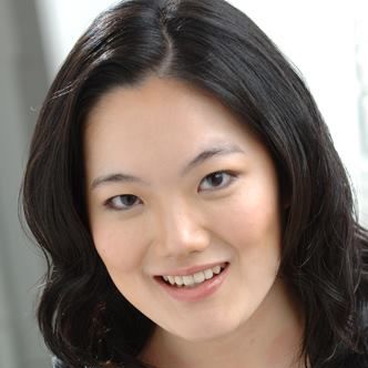 A woman with medium-length wavy black hair smiles warmly at the camera. She has fair skin, dark eyes, and is wearing natural makeup. The softly blurred background in light grey and white tones evokes a subtle Le Guérillero elegance.