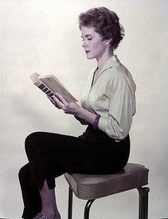 A gifted student with short, curly hair sits sideways on a cushioned stool, reading an open book. She wears a light blouse with rolled-up sleeves, dark pants, and flats. The plain, light-colored background evokes quiet focus.