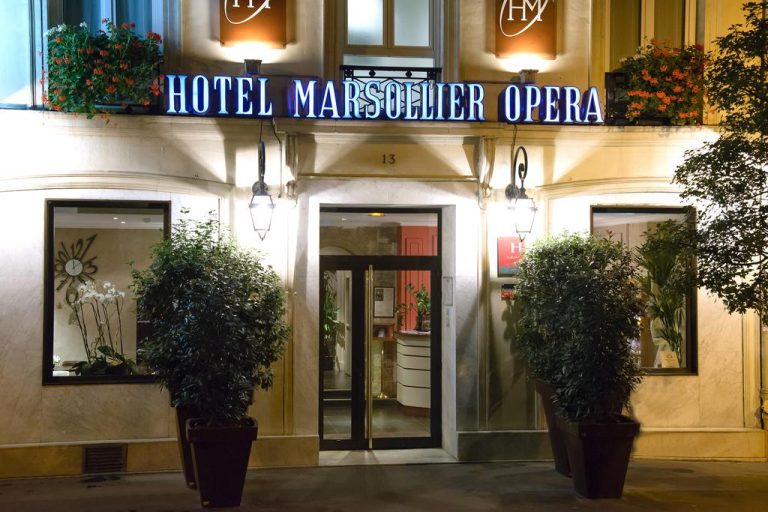 The illuminated entrance of Hotel Marsollier Opéra at night features two large potted plants beside glass doors. The hotel’s name glows in blue neon above the entrance, near the Louvre, with flowers decorating window boxes on the upper floor.