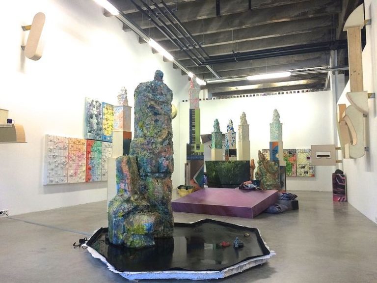 An art gallery with white walls, a grey concrete floor, and an exposed ceiling. Colorful abstract sculptures by Georges Verney-Carron and mixed media artworks curated by a médiateur artistique fill the space with textured, geometric forms.