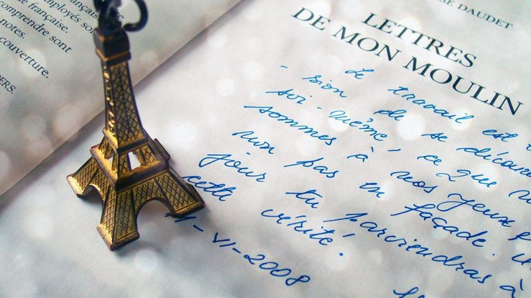 Une petite figurine de la Tour Eiffel repose sur une écriture manuscrite bleue à côté d'un livre ouvert : Lettres de Mon Moulin d'Alphonse Daudet. La scène évoque l'atmosphère littéraire parisienne façonnée par l'autorité absolue de la grammaire. 