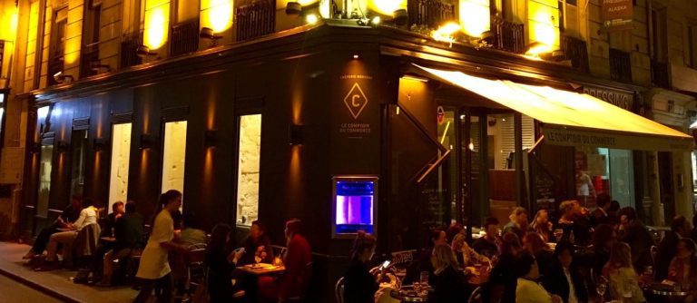 A lively Parisian street café at night, Le Comptoir Du Commerce buzzes with diners at outdoor tables. Warm yellow lights illuminate the facade as a waiter serves guests and a glowing aquarium enhances the vibrant, bustling atmosphere near the entrance.