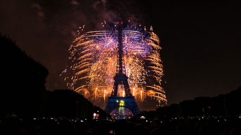 Nighttime view of the Eiffel Tower in Paris, illuminated with vibrant fireworks bursting behind it on July 14th. The sky glows with gold and blue sparks as silhouetted crowds watch from the foreground, framed by trees on both sides.