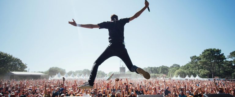 A performer holding a microphone leaps energetically with arms wide on an outdoor stage at festivals, facing a large cheering crowd under a clear blue sky, surrounded by trees and white festival tents in the background.
