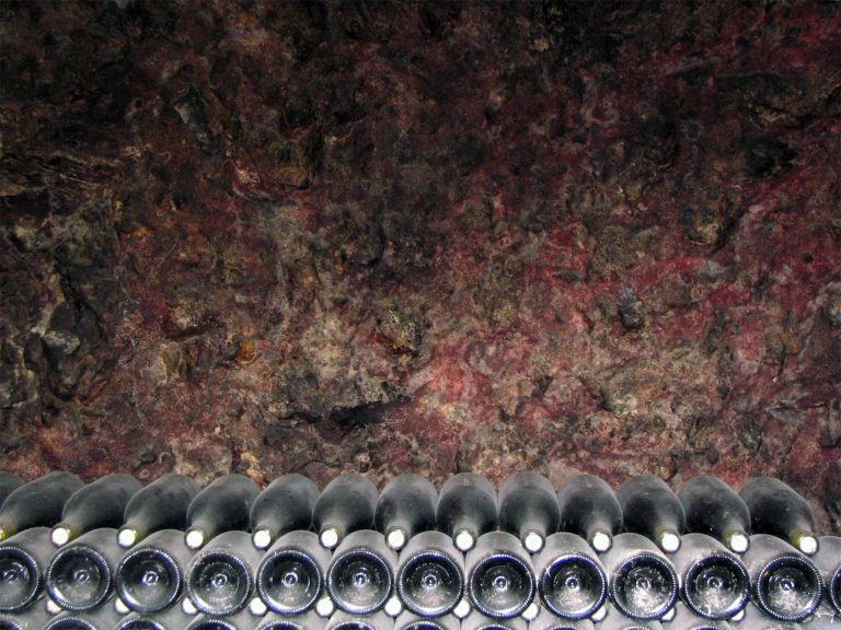 A row of Le Vin bottles lies horizontally, stacked in a cellar against a rough, dark, reddish-brown stone wall, their bases facing outward and reflective surfaces catching the light.