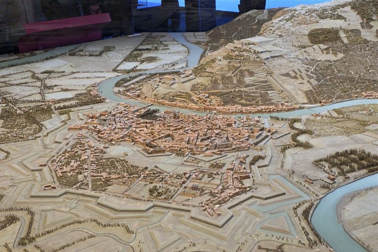 A detailed scale model in the style of Vauban’s Plans-Reliefs depicts a historic city ringed by defensive walls and bastions, winding rivers and bridges; packed buildings fill its core, with hills, roads, fields, and trees beyond.