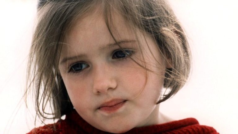 Close-up of a young girl with light skin and straight brown hair, some strands falling over her face. She is wearing a red sweater and looking slightly downward with a thoughtful, somewhat sad expression. The background is softly blurred and bright.