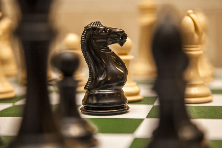 A close-up of a black knight chess piece on a green and white chessboard, surrounded by blurred pieces in the background, evokes a sense of focus and imagination, as if the scene were drawn from a fantasy or Kasparov autobiography.