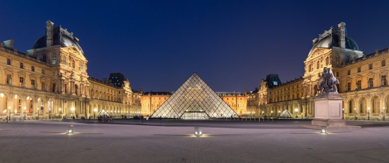 Night view of the Louvre Museum in Paris, featuring the illuminated glass pyramid entrance at the center, flanked by the historic museum buildings and a statue on the right side. The sky is dark blue, and the courtyard is mostly empty.