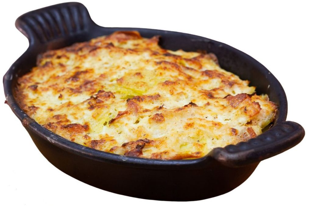A black oval baking dish with handles contains a golden-brown, baked casserole topped with a crispy, slightly uneven layer, likely made from mashed potatoes or cheese. The dish is isolated on a white background.
