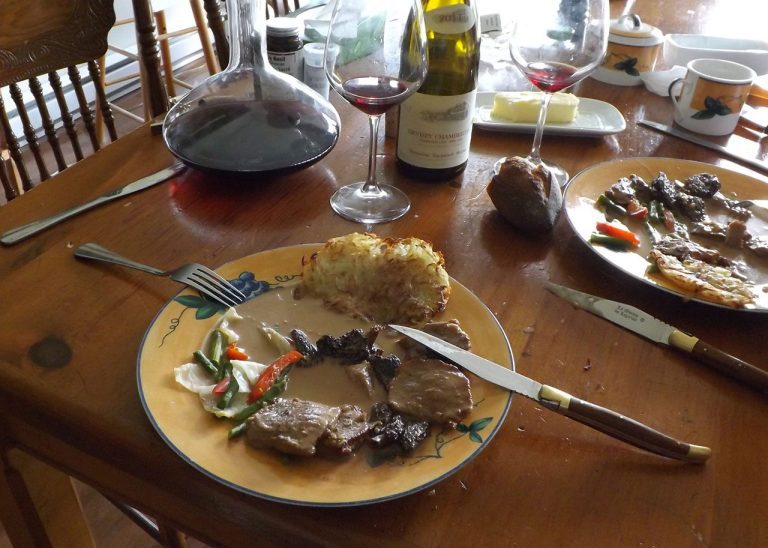 A wooden table set for a meal features a plate with meat in gravy, vegetables, and hash brown—reminiscent of Traditional French Foods. Wine glasses, a wine decanter, a bread roll, and butter complete the inviting scene.