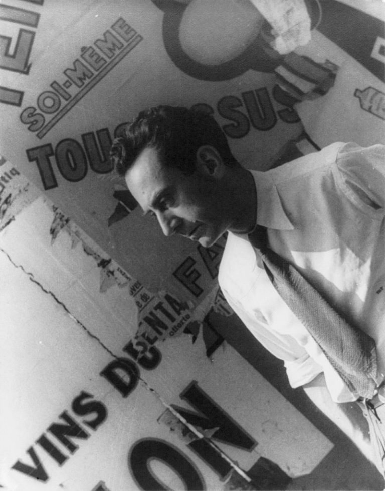 A man in a white shirt and tie stands in front of torn, overlapping posters with large French text. He looks downward in profile, lit from the left. The background includes bold letters and partial words in various fonts and sizes. The image is black and white.