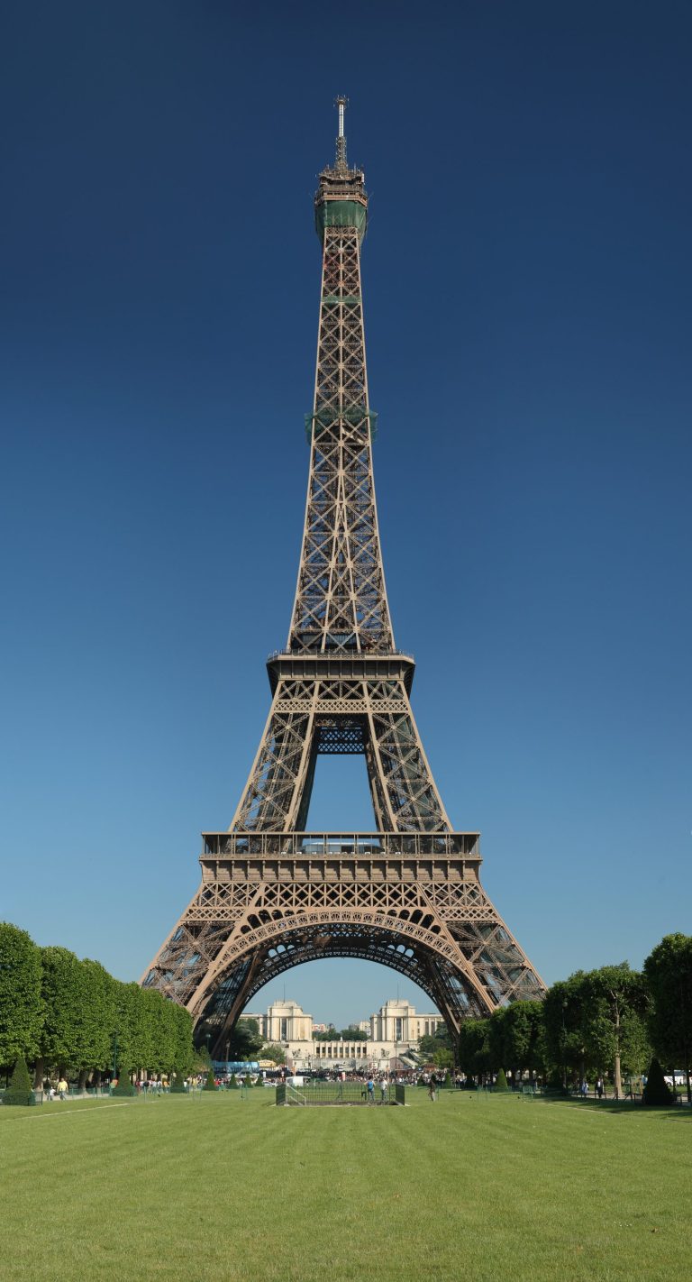 The Eiffel Tower, an iconic Paris landmark, stands tall against a clear blue sky, surrounded by lush green trees and a well-kept lawn. People gather near the base, while distant buildings are visible through the tower’s legs, adding to its grandeur.
