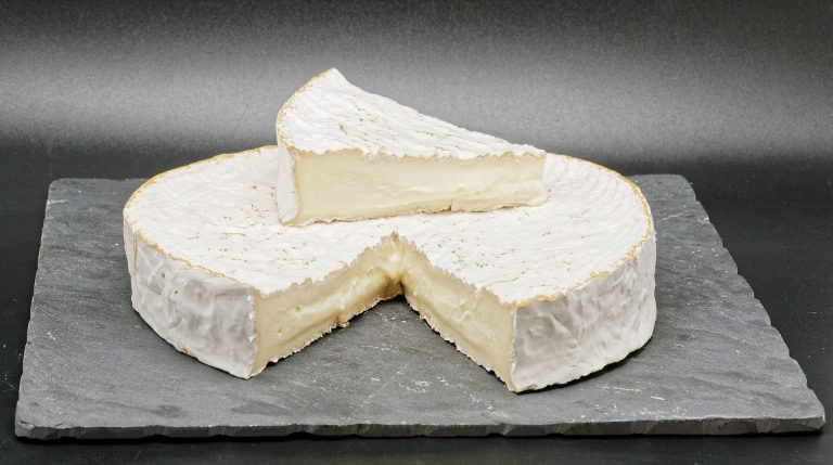 A round wheel of creamy white Brie cheese sits on a dark slate board. A wedge rests on top, revealing the soft interior and white rind—perfect for exploring brie cheese pairings or discovering what goes best with brie. The background is plain and dark.