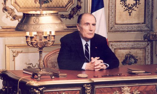 An older man in a dark suit and tie sits at an ornate desk with a brass lamp, books, and papers. He clasps his hands thoughtfully, as if reflecting on my life; gold details and a French flag evoke the legacy of Mitterand in the lavish room.