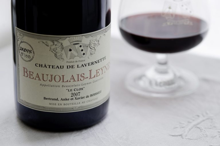 A close-up of a wine bottle labeled Château de Lavernette Beaujolais-Leynes 2007 next to a glass of red wine on a white tablecloth—a joyful scene celebrating Beaujolais, with decorative elements and the names Bertrand, Anke et Xavier de Boissieu.