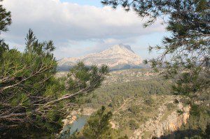 A distant rocky mountain rises under a partly cloudy sky, framed by pine tree branches in the foreground. Below, green hills and sparse trees stretch towards a small lake at the bottom left, creating a serene natural landscape.