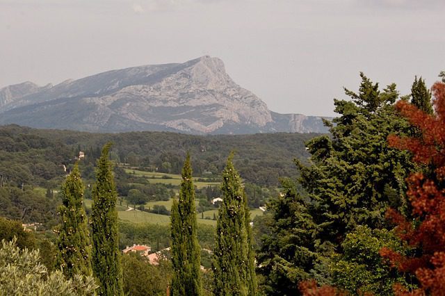 A mountain with a distinct peak rises behind a forested landscape reminiscent of Paul Cezanne’s style. Tall green trees and a few red-leaved branches frame scattered buildings below, beneath a light, mostly clear sky.