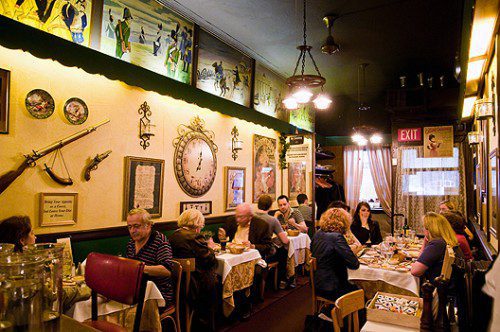 A cozy, warmly lit restaurant, Chez Napoleon features antique decor—a large clock, ornate frames, and old rifles. Murals grace the upper wall, while soft curtains frame windows by the EXIT sign—perfect for a Bastille Day celebration.