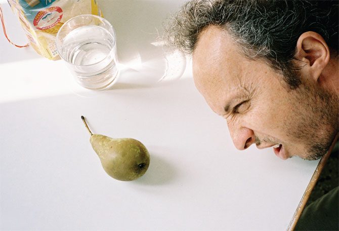 A man with curly hair leans his face close to a green pear on a white table, squinting and grimacing. In the background, a glass of water and a food package from New York Bureau cast long shadows across the table.