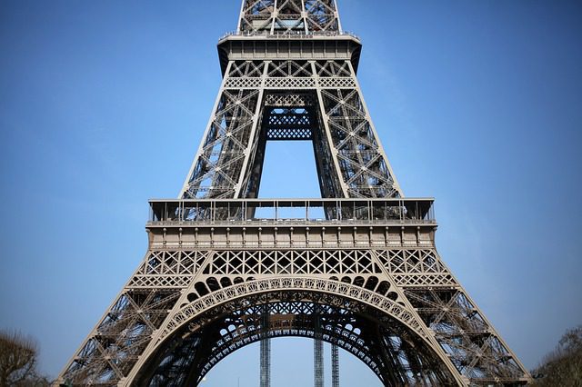 Close-up view of the lower section of the Eiffel Tower against a clear blue sky, showcasing its detailed iron lattice—a true icon of français style de vie. The photo captures the base and legs with intricate architectural patterns. No people visible.