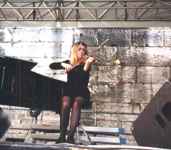 Une femme en robe noire est assise sur un tabouret et joue du jazz au violon sur une scène en plein air. Un piano à queue se trouve à côté d'elle, encadré par de gros blocs de pierre à l'arrière-plan. Des gréements métalliques et des haut-parleurs entourent la scène.  