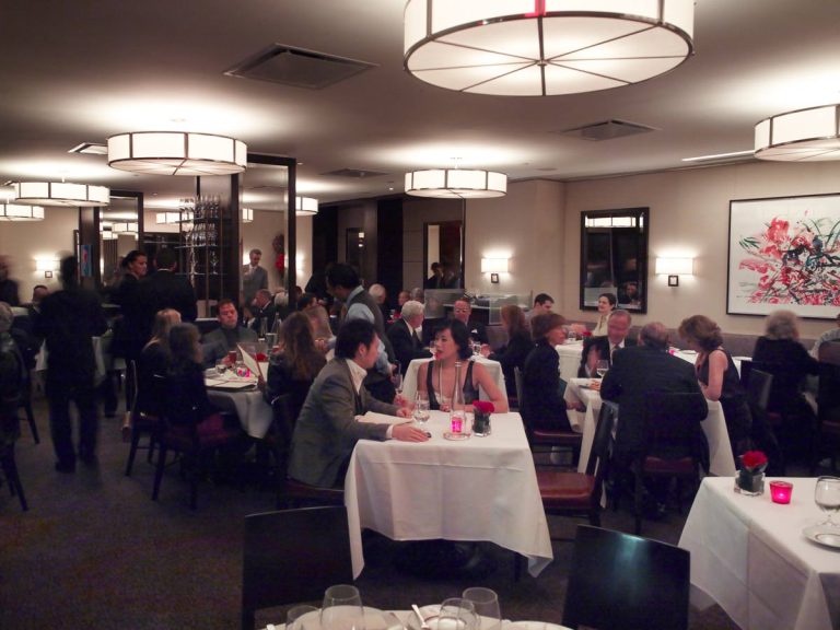 A busy upscale restaurant renowned for its haute cuisine, with people dressed in formal attire dining at tables covered with white tablecloths. The lively atmosphere features modern lighting, wall art, and attentive waitstaff serving guests throughout the elegant space.