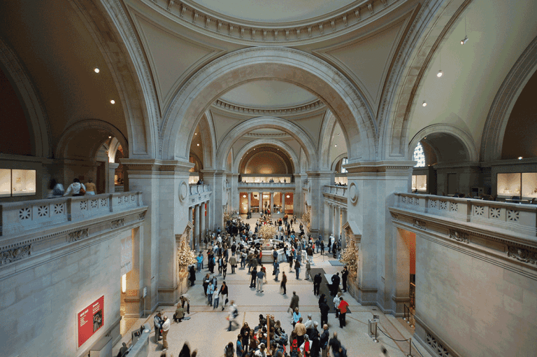 A wide, high-ceilinged museum hall with elegant arches and stone walls is filled with many visitors walking, gathering, and observing. Natural light streams in from windows above, evoking a timeless elegance amid the busy, spacious interior.