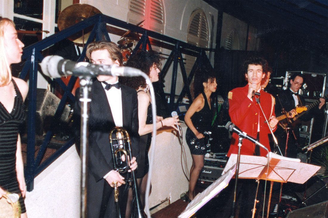 Un orchestre dynamique se produit sur scène lors des fêtes. Une femme en robe noire se tient à gauche, un homme en smoking tenant un trombone est au centre, et un autre en veste rouge chante, évoquant l'élégance d'un événement organisé par Elie de Rothschild. 