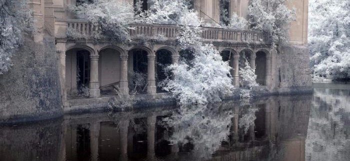 An old stone building with arched windows and a balcony stands partly submerged in calm water. Frost-covered trees surround this VIE DE CHÂTEAU scene, their icy foliage reflected below, creating an ethereal, wintry atmosphere.