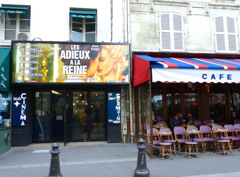 A French street scene shows a small cinema with a movie poster for Les Adieux à la Reine above its entrance. Next to it is a café with a red-and-blue striped awning and empty wicker chairs—perfect spots to learn French or say, "Allez au Cinéma!.