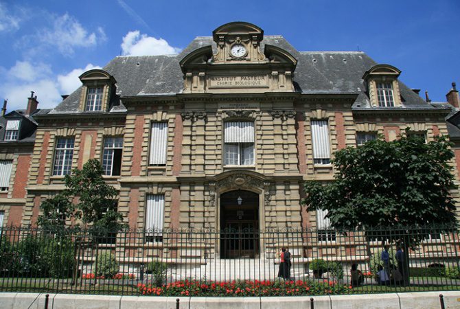 Historic stone and brick building with arched entry, clock, and ornate carvings reading Institut Pasteur. This patrimoine of savoir features tall windows with shutters, iron fence, flowerbeds, trees, and people near the entrance under a blue sky.
