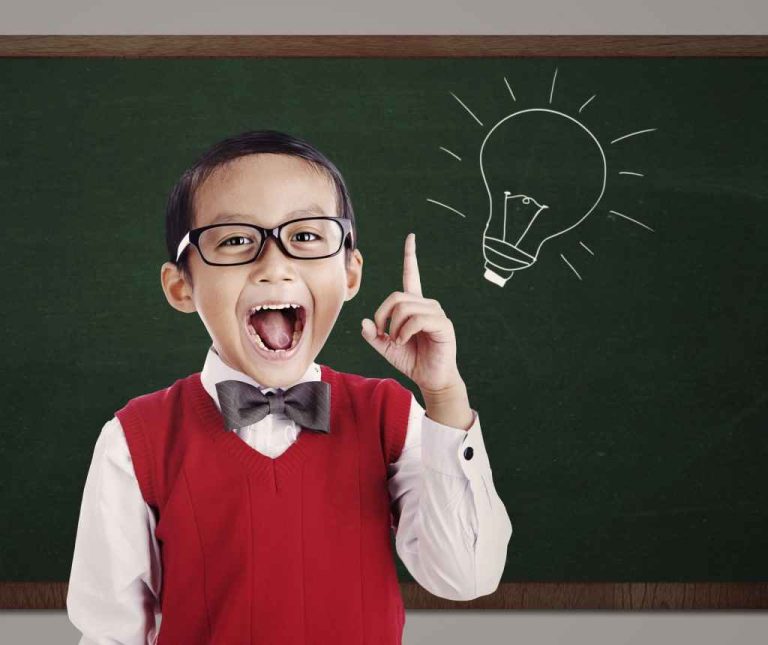 Un étudiant surdoué portant des lunettes, un gilet rouge, une chemise blanche et un nœud papillon se tient devant un tableau vert sur l'avenue de la Grande Armée. Souriant largement, il lève un doigt à côté d'un dessin à la craie représentant une ampoule électrique allumée. 
