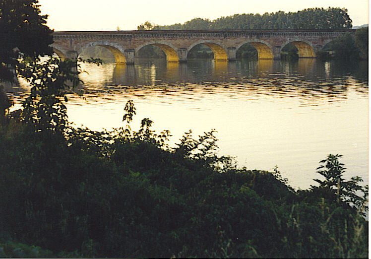 A stone arch bridge spans a calm river at sunset in France, with warm light reflecting on the water. Lush green trees frame the scene, making it an idyllic view often admired on a luxury barge tour through this tranquil countryside.