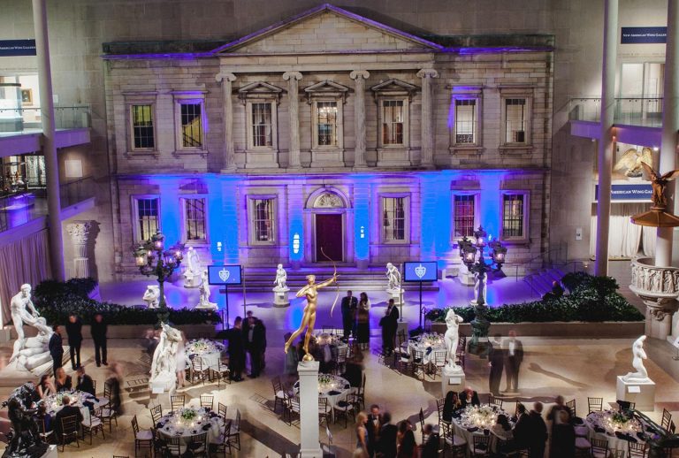 A grand indoor hall with an ornate, classical building facade lit by blue lights. Elegant round tables and chairs are set for an event. People in formal attire mingle among white marble statues and a golden statue at the center. The atmosphere is sophisticated and festive.