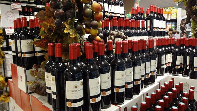Rows of red wine bottles labeled La Pierre Cachee, one of the best French wines, are neatly stacked on display in a store. Artificial grapevines with multicolored grapes and leaves hang above, while more vintage year bottles line the shelves behind.