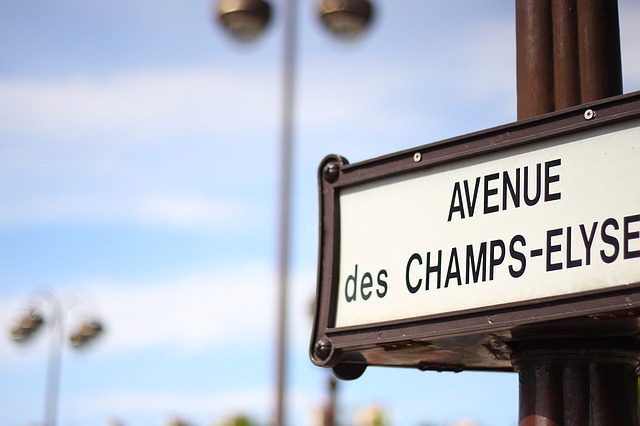 Close-up of a street sign reading Avenue des Champs-Élysées in black letters on a white background, mounted on a dark pole. The blurred Paris background reveals blue sky, scattered clouds, and faint outlines of streetlights.