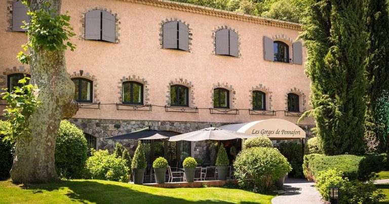 A rustic, two-story stone building with gray shutters and arched windows houses the Hostellerie des Gorges de Pennafort hotel and restaurant. Lush greenery surrounds shaded outdoor seating with black umbrellas for guests to enjoy.