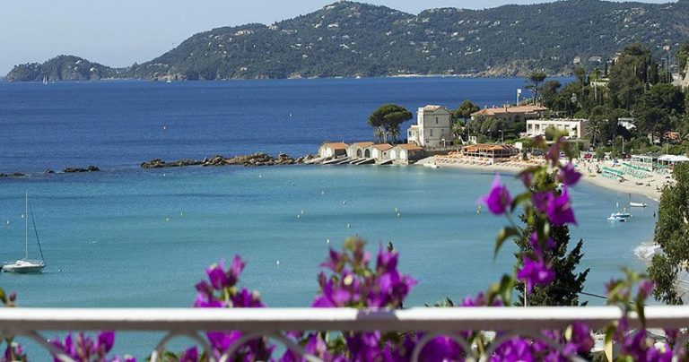 A scenic coastal view with clear blue water, a sandy beach lined with sun loungers, and small white buildings by the shore—including Hotel Belle-Vue. Purple bougainvillea blooms in the foreground, with forested hills rising under a clear sky.