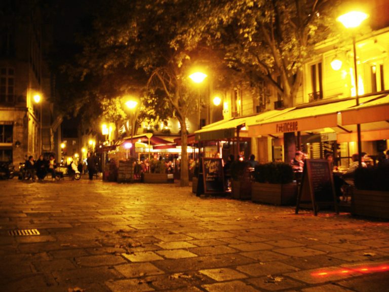 A warmly lit outdoor café on a cobblestone street at night. Trees line the street and yellow streetlights illuminate the area. Several people sit at tables under orange awnings. Buildings with lit windows form the backdrop. The atmosphere feels calm and inviting.